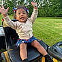 arms_raised, black_shoes, child, grass, greenery, hair_bows, happy, jacket, lawnmower, nature, outdoor, park, person, playful, seat, skirt, smiling, sunlight, toddler, tree