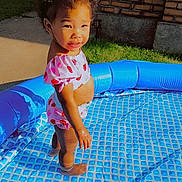 Kaylani is registered to the contest to win money with this photo: anklet, brick_wall, child, concrete, curly_hair, cute, daytime, grass, happy, inflatable_pool, outdoor, play, side_view, smiling, summer, sunlight, swimsuit, toddler, truck, water
