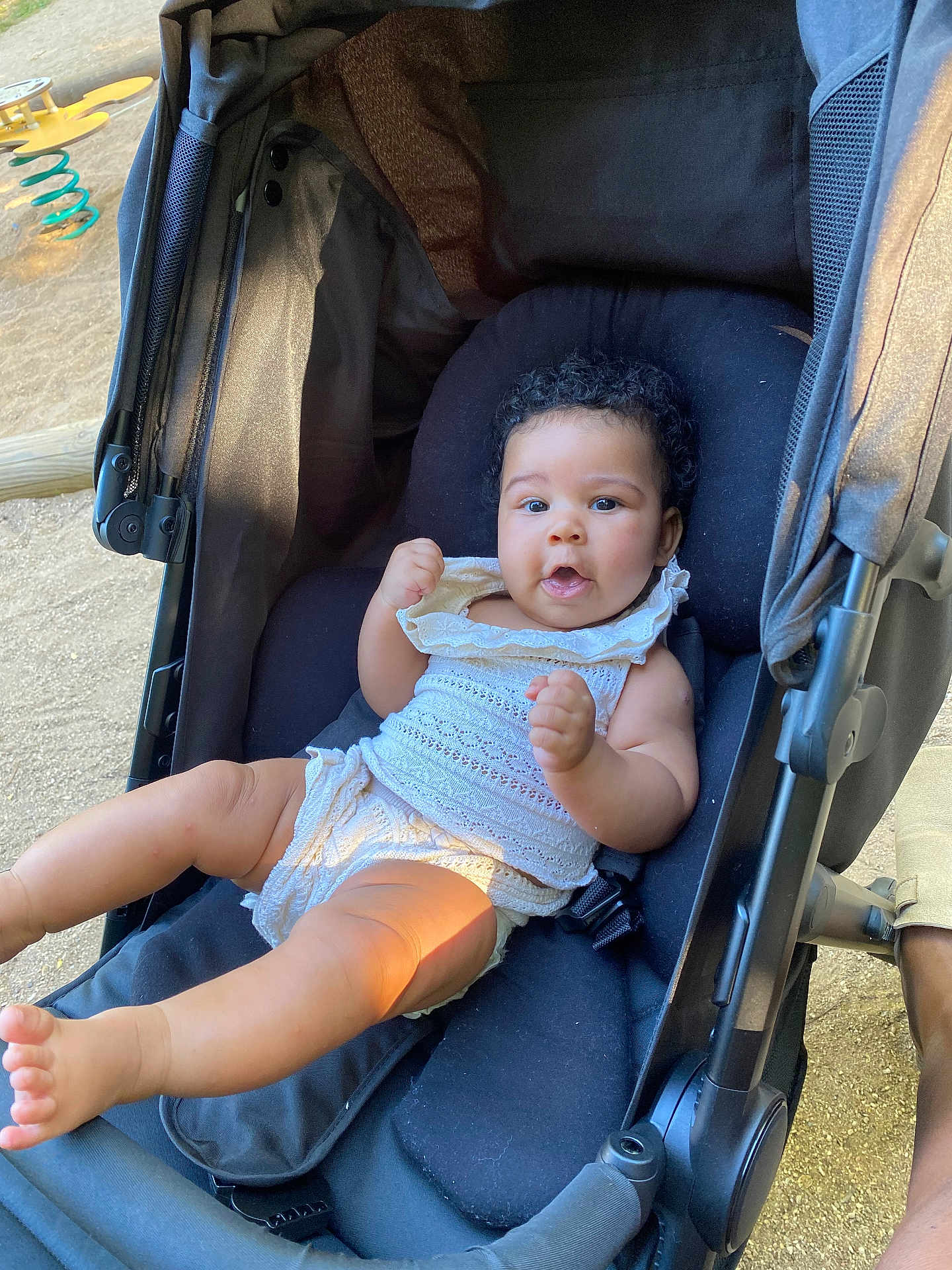 Lenaëlle participe au concours pour gagner de l'argent avec cette photo : baby, child, stroller, curly_hair, white_outfit, outdoor, playful, legs, face, expression, seat, comfort, sunlight, person, infant, cute, summer, relaxing, hands, young