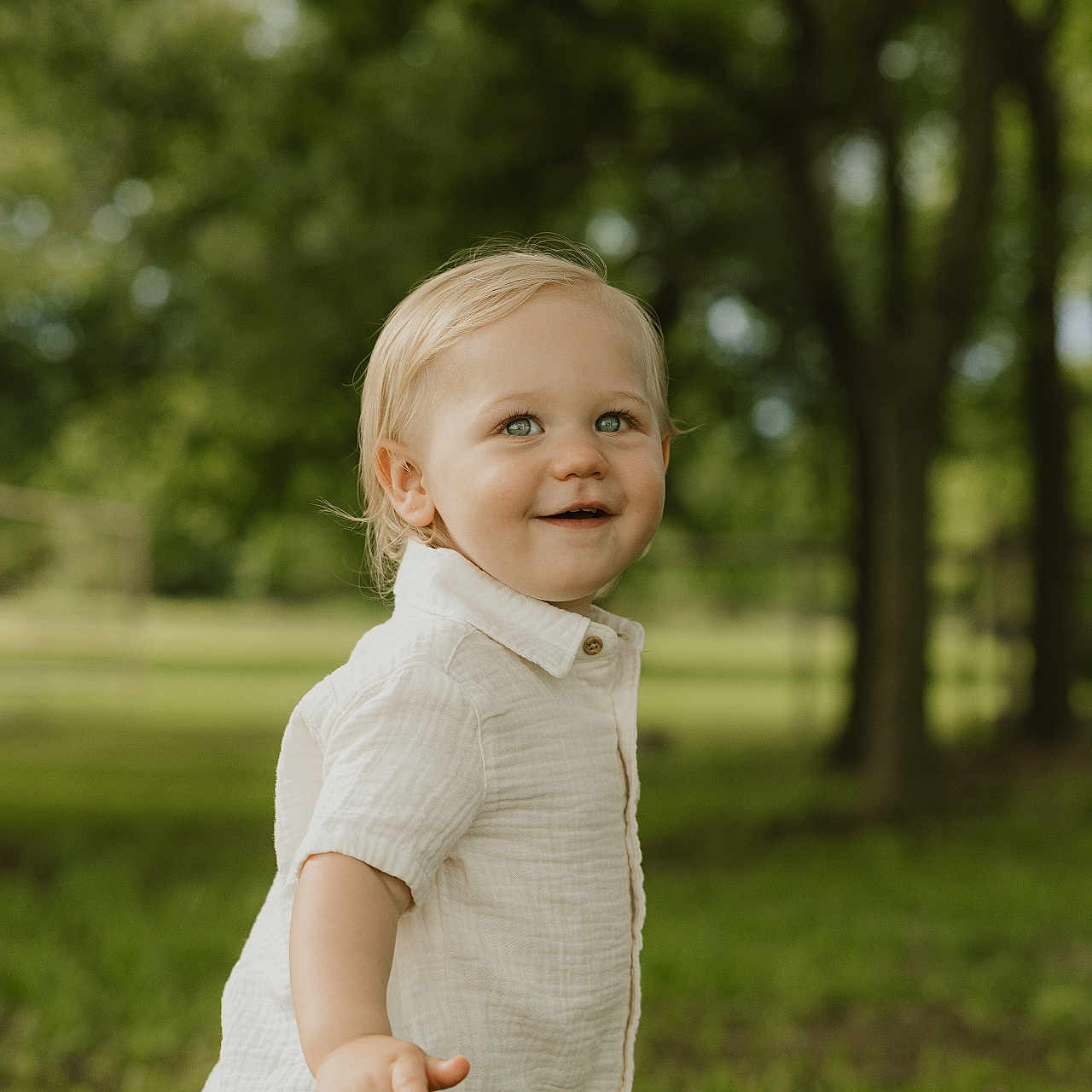 Carson is registered to the contest to win money with this photo: background_blur, blond_hair, boy, casual_clothing, child, cute, daylight, grass, green_eyes, happy, nature, outdoor, pointing, portrait, shirt, shorts, smiling, toddler, trees, young_child