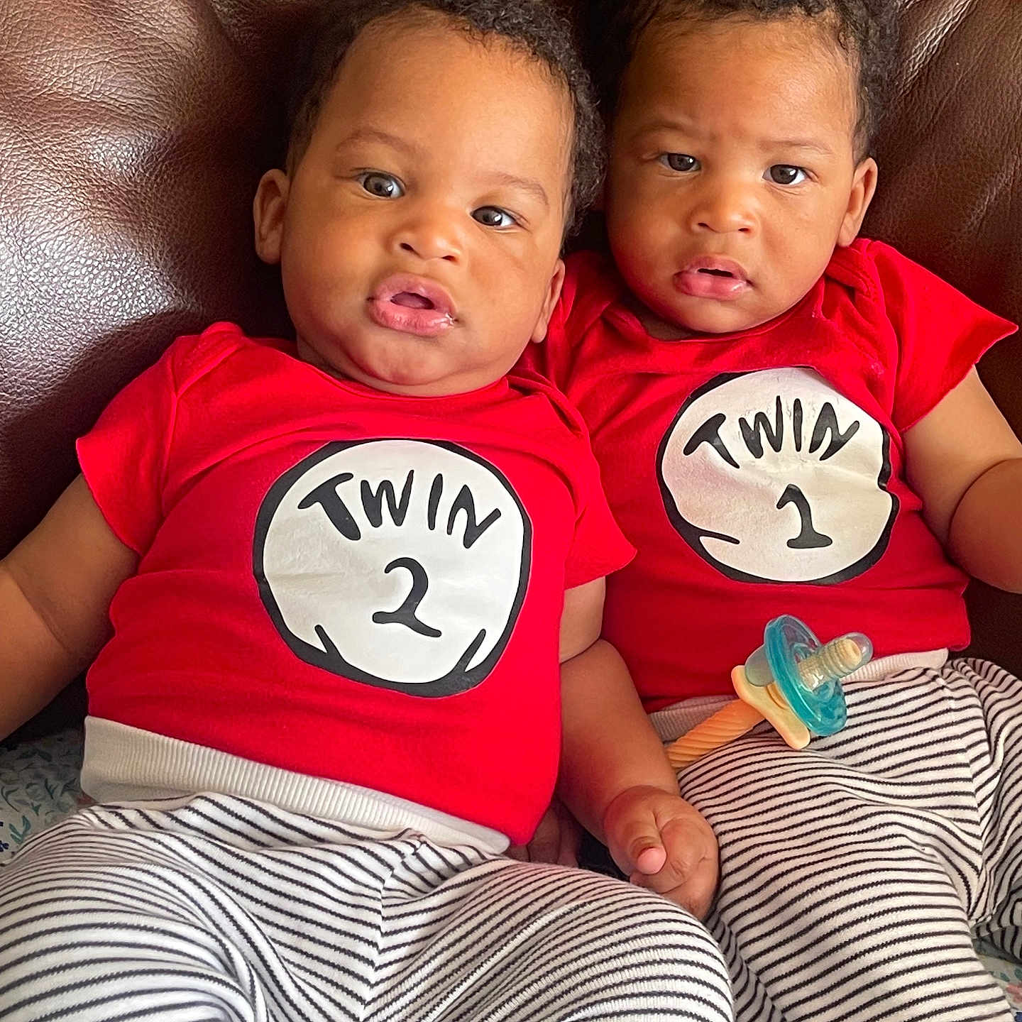 Zamir Jahziel joined the competition — help win amazing prizes! twin, baby, toddler, red_shirt, striped_pants, pacifier, leather_couch, sitting, indoor, child, cute, matching_clothes, siblings, infant, person, face, expression, curious, close, bonding
