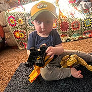 Judson is registered to the contest to win money with this photo: toddler, child, toy, construction_vehicle, action_figure, yellow_cap, crocheted_blanket, couch, rug, carpet, indoor, play, sitting, foot, clothing, person, sunlight, living_room, home, casual