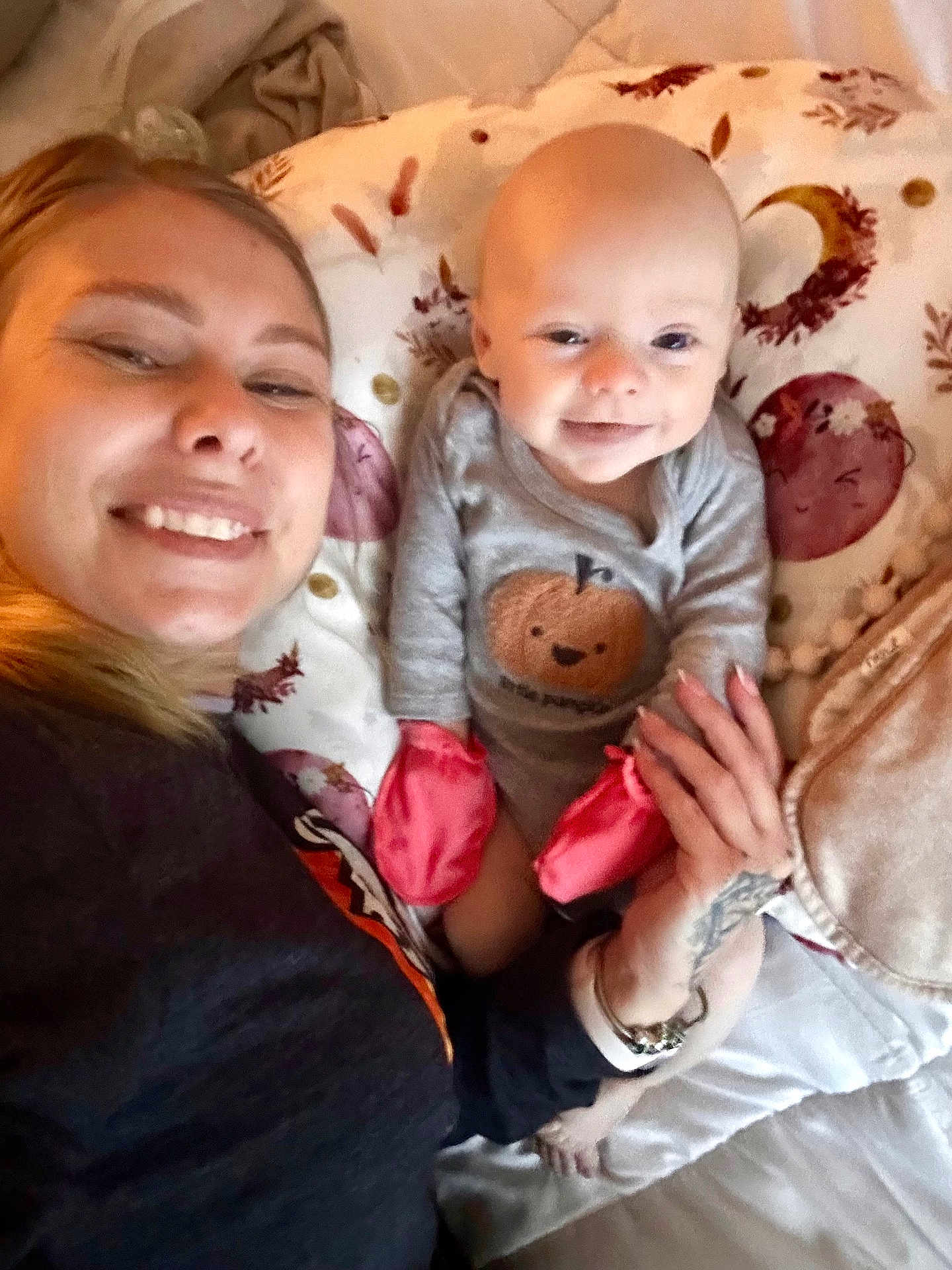 Monroe is registered to the contest to win money with this photo: woman, baby, smiling, selfie, indoor, blanket, pillow, mittens, onesie, face, hand, tattoo, happy, lying_down, cozy, bed, person, cute, infant, casual