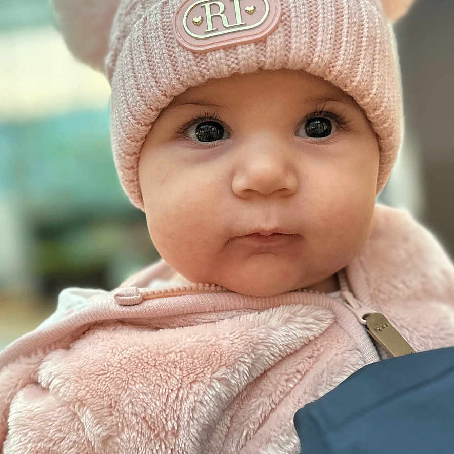 Celine is registered to the contest to win money with this photo: adorable, baby, child, close_up, cute, face, fuzzy_clothing, hat, indoor, infant, jacket, large_eyes, person, pink, pom_poms, portrait, soft_texture, warm_clothing, winter_clothing, young_child