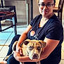 woman, dog, indoor, glasses, black_shirt, floral_pattern, tile_floor, seated, brown_dog, blue_eyes, furniture, table, chair, pet, casual, portrait, home, floor, cute, human_animal_interaction