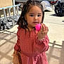 braided_hair, casual_clothing, child, concrete, curly_hair, daylight, earrings, girl, holding_object, outdoor, person, pink_dress, plastic_bag, plastic_egg, playful, portrait, shadows, small_child, sunlight, young_child