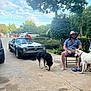 man, dog, black_dog, white_dog, car, classic_car, chair, outdoor, driveway, trees, grass, sky, clouds, person, footwear, hat, beard, can, conversation, nature