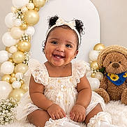 Slaia is registered to the contest to win money with this photo: baby, child, infant, smiling, portrait, sitting, dress, headband, bow, teddy_bear, stuffed_animal, balloons, gold, white, celebration, birthday, flowers, sunflower, indoors, photography