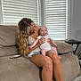 baby, woman, couch, indoor, blinds, smile, tongue_out, holding, happy, blond_hair, shorts, toddler, casual_clothing, wooden_floor, stool, phone, home, light, person, relaxing