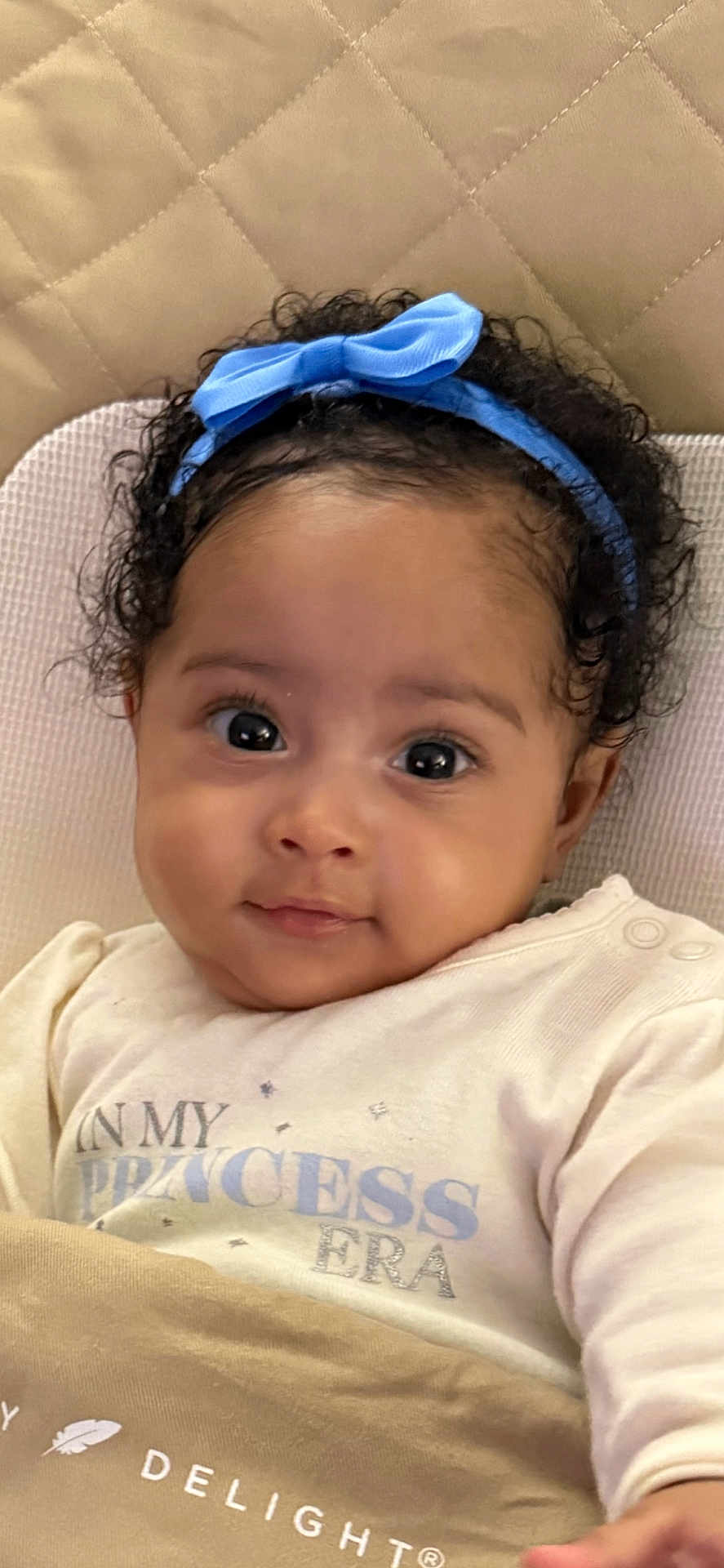 Brylee joined the competition — help win amazing prizes! baby, infant, headband, blue_bow, big_eyes, curly_hair, onesie, princess_text, blanket, quilted_background, portrait, closeup, cheeks, eyelashes, smile, bedding, cute, human_face, sitting, soft_lighting