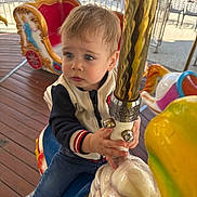 Timéo participe au concours pour gagner de l'argent avec cette photo : child, toddler, carousel, horse, amusement_ride, golden_pole, outdoor, wooden_floor, colorful, playful, clothing, jacket, jeans, sneakers, daylight, expression, hands, face, eyes, fun