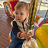 child, toddler, carousel, horse, amusement_ride, golden_pole, outdoor, wooden_floor, colorful, playful, clothing, jacket, jeans, sneakers, daylight, expression, hands, face, eyes, fun