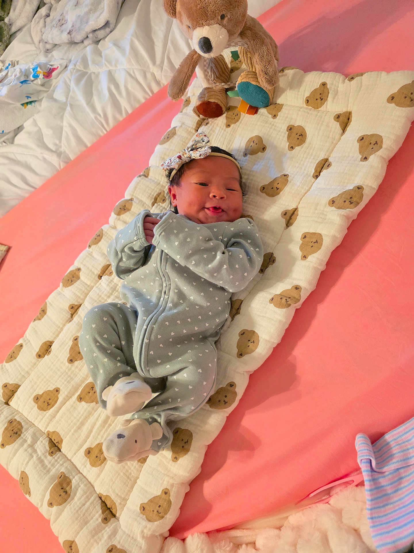 Gia is registered to the contest to win money with this photo: baby, infant, onesie, blanket, teddy_bear, plush_toy, bed, sleepwear, headband, smiling, cute, cozy, child, soft_toy, pink, fabric, pattern, lying_down, indoors, resting