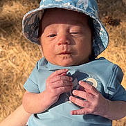 Khalil is registered to the contest to win money with this photo: baby, infant, child, blue_clothing, bucket_hat, outdoor, sunlight, hands, face, expression, summer, nature, pine_needles, casual, cute, small_hands, person, young, portrait, daylight
