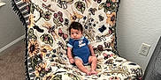 Khalil joined the competition — help win amazing prizes! baby, blanket, blue_clothing, butterflies, carpet, chair, child, cozy, curious_expression, electrical_outlet, floral_pattern, furniture, indoor, infant, onesie, person, seated, sunflowers, text_on_clothing, wall