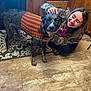 dog, person, sweater, indoor, kitchen, floor, tile, rug, wood_cabinet, smiling, petting, animal, canine, happy, clothing, casual, home, companion, friendship, brown