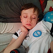Esteban a rejoint le concours — aidez-le/la à gagner de superbes lots ! child, sleeping, bed, pillow, blanket, white_shirt, emblem, hand, face, resting, closed_eyes, indoor, young_boy, hair, comfort, relaxation, casual_clothing, portrait, person, tired
