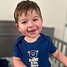 toddler, child, smile, happy, blue_tshirt, text, indoors, bedroom, crib, short_hair, face, person, cute, portrait, young_child, casual_clothing, baby, joyful, expression, background_blur