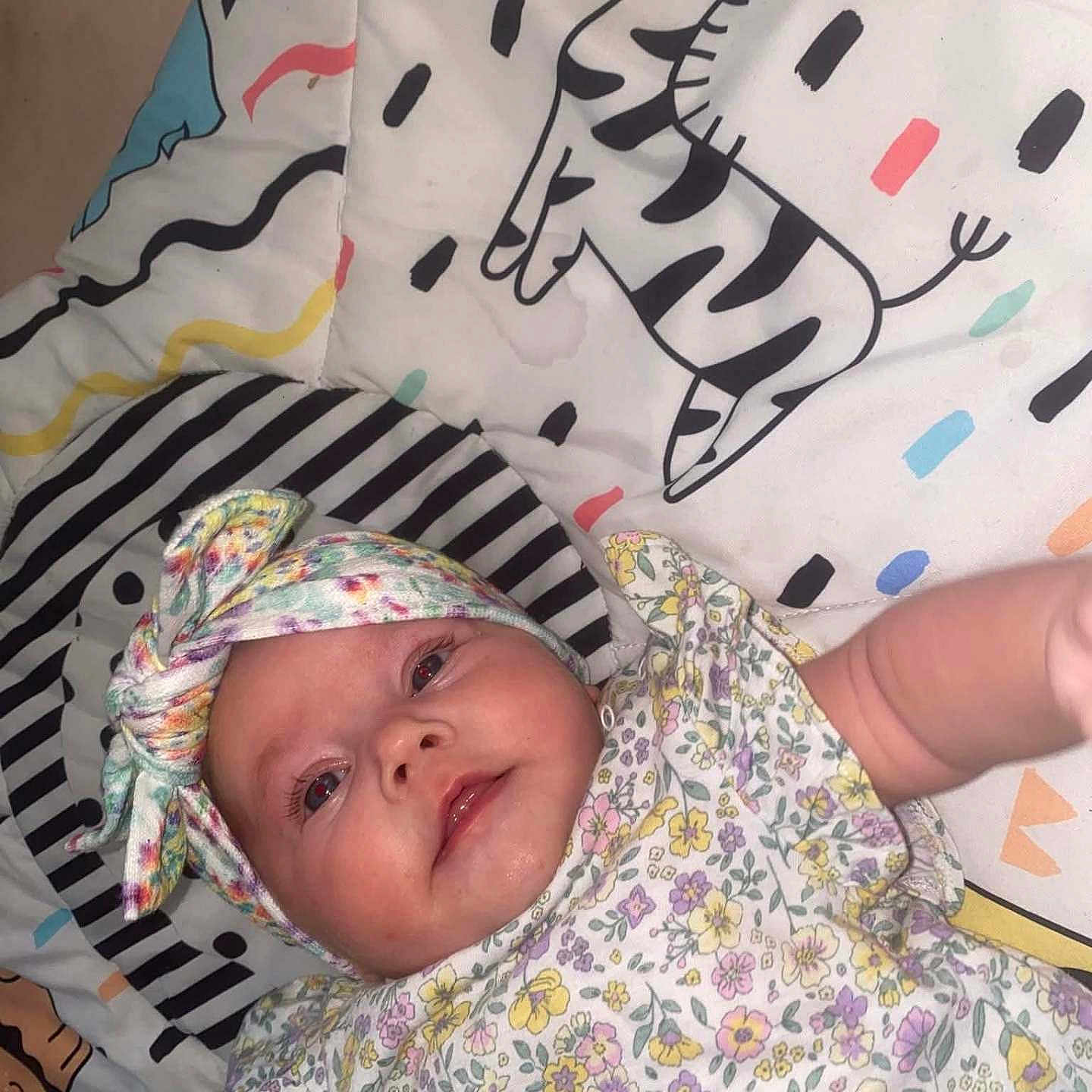 Maeve joined the competition — help win amazing prizes! baby, blanket, cartoon, child, closeup, colorful, cute, face, floral, headband, indoor, infant, lying_down, patterned_clothing, playful, reaching, smiling, soft_toys, young_child, zebra