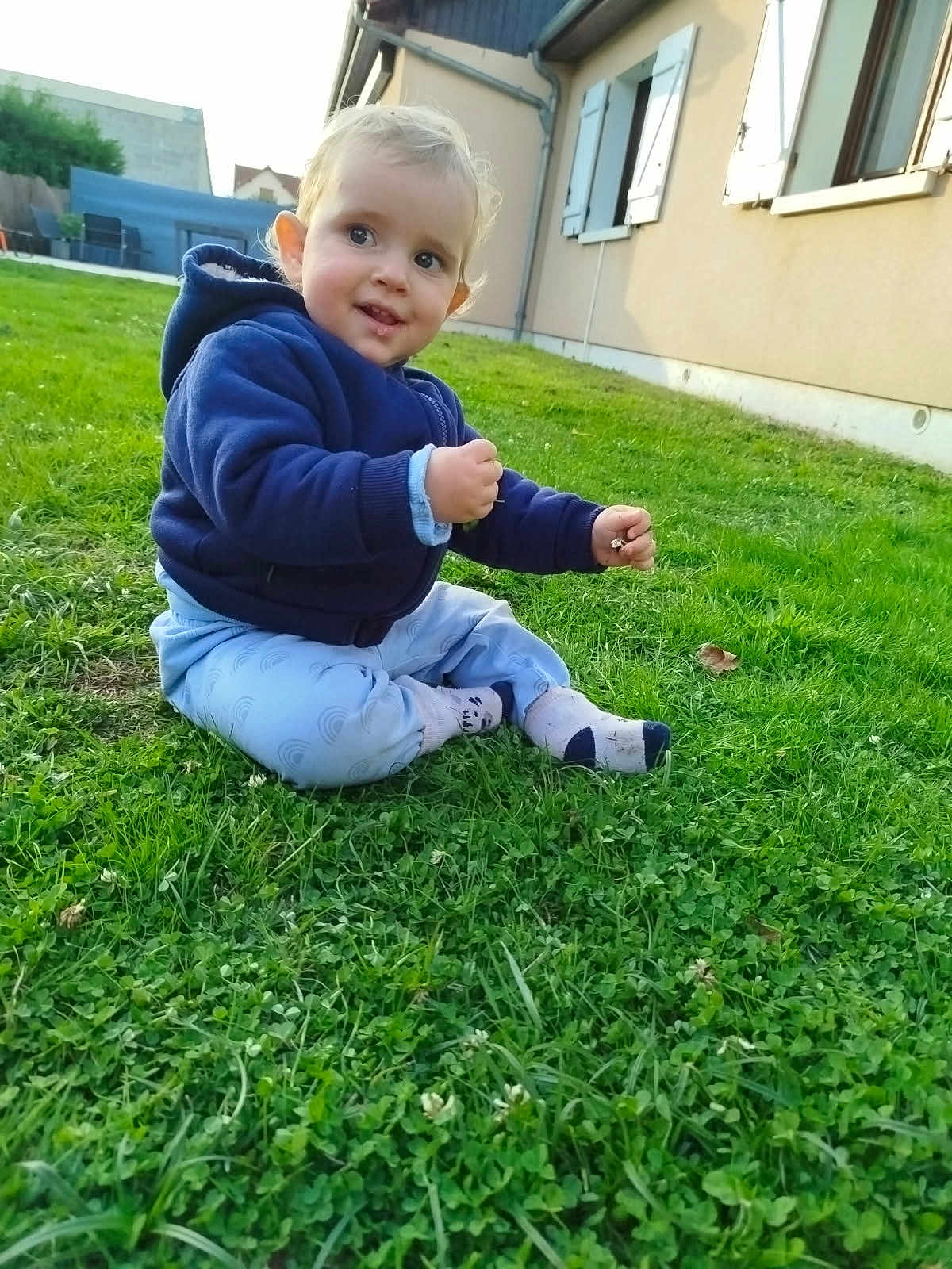 Rafael a rejoint le concours — aidez-le/la à gagner de superbes lots ! toddler, child, grass, outdoor, smile, blue_hoodie, pants, socks, house, window, shutters, greenery, nature, young_child, baby, sitting, curious, daylight, casual_clothing, suburb
