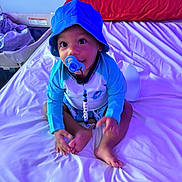 Tyrell is registered to the contest to win money with this photo: baby, child, pacifier, hat, blue_clothing, bed, pillow, indoor, sitting, cute, infant, person, face, skin, feet, hands, long_sleeve, white_bed_sheet, red_pillow, looking_up