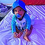 baby, child, pacifier, hat, blue_clothing, bed, pillow, indoor, sitting, cute, infant, person, face, skin, feet, hands, long_sleeve, white_bed_sheet, red_pillow, looking_up