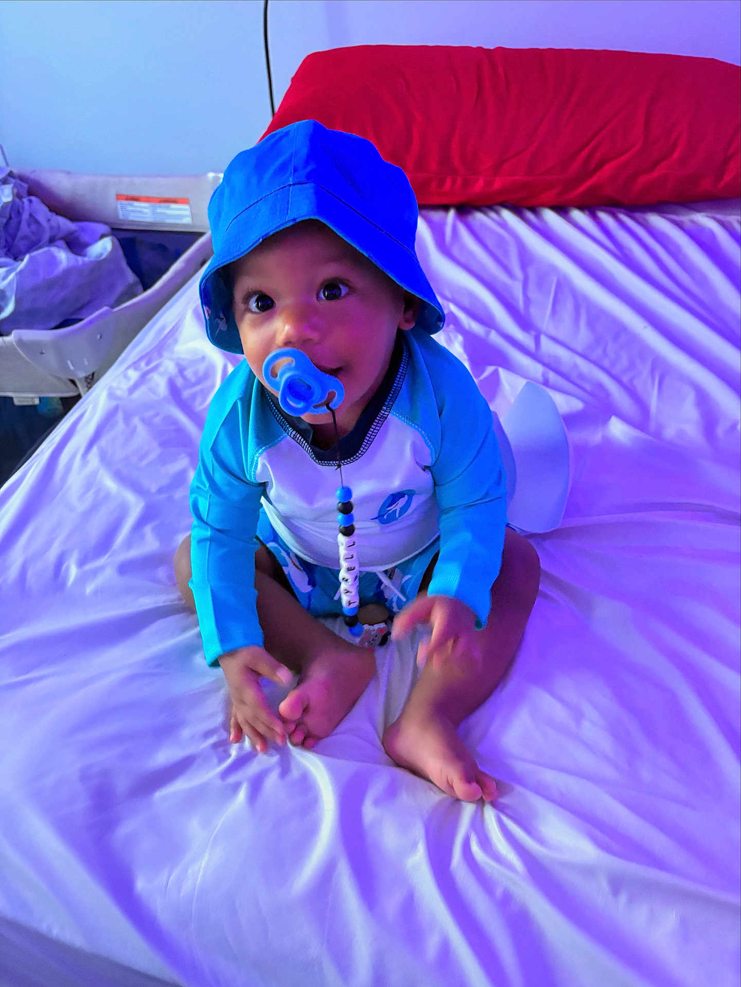Tyrell is registered to the contest to win money with this photo: baby, child, pacifier, hat, blue_clothing, bed, pillow, indoor, sitting, cute, infant, person, face, skin, feet, hands, long_sleeve, white_bed_sheet, red_pillow, looking_up