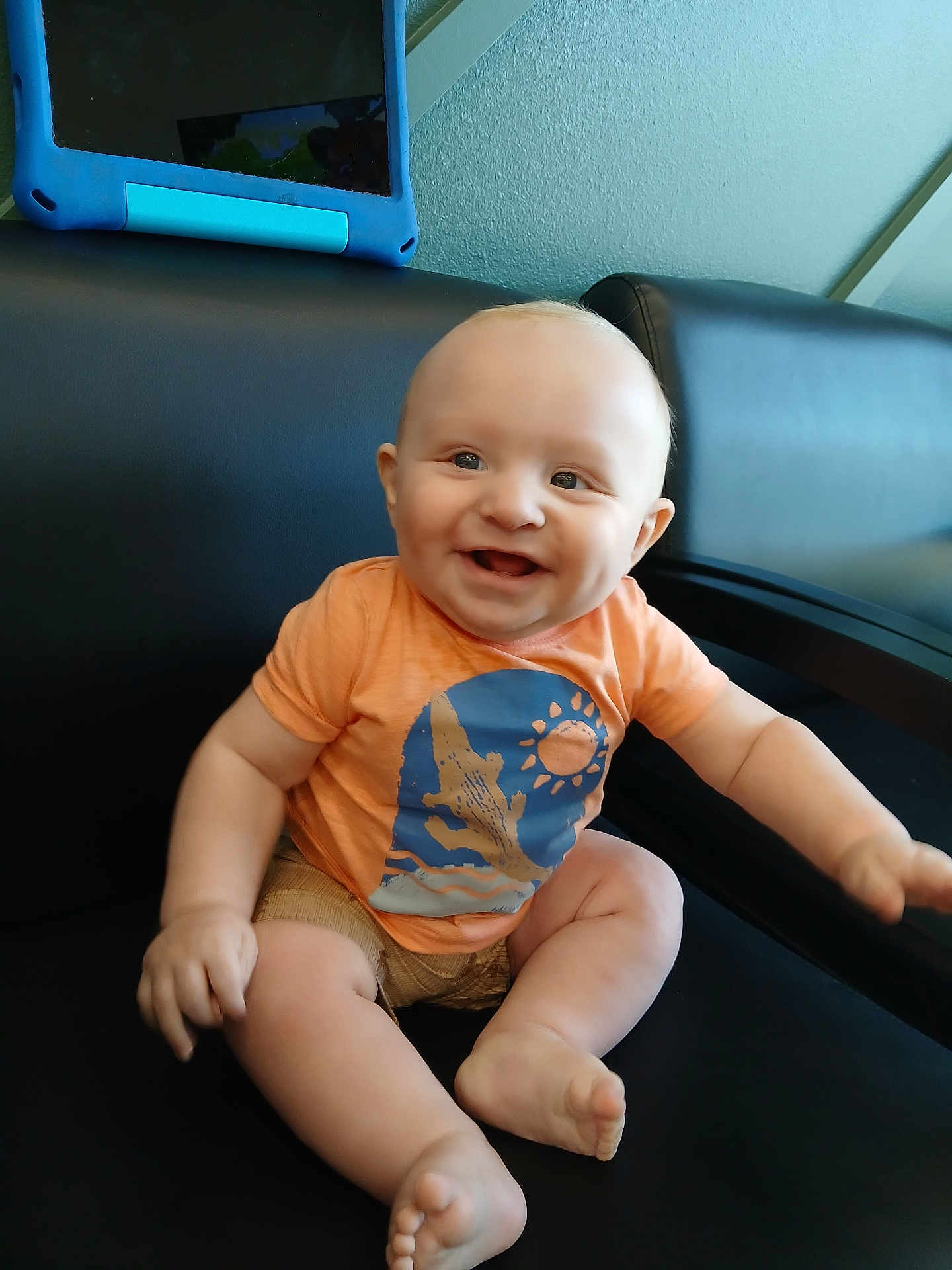 Archer is registered to the contest to win money with this photo: baby, child, smiling, orange_shirt, sitting, black_chair, tablet, indoor, happy, cute, skin, head, legs, feet, arms, cactus_design, sun_design, person, infant, casual
