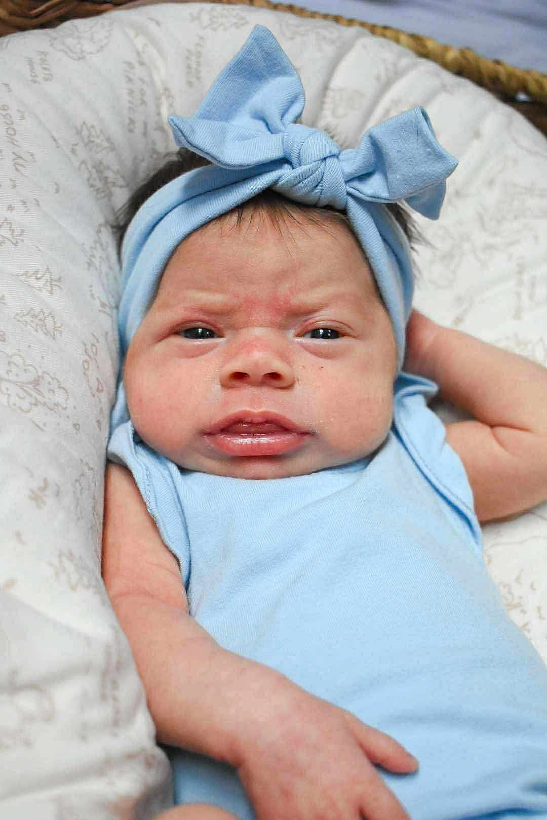 Veronica is registered to the contest to win money with this photo: baby, infant, headband, blue, bow, face, expression, grumpy, clothing, cushion, pattern, skin, person, newborn, lying_down, closeup, indoors, cute, child, portrait