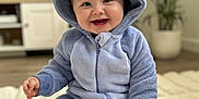 Alfie joined the competition — help win amazing prizes! baby, child, smiling, blue_onesie, hood, plush_clothing, sitting, indoor, living_room, rug, plant, cabinet, television, happy, cute, infant, portrait, cozy, soft_texture, playful