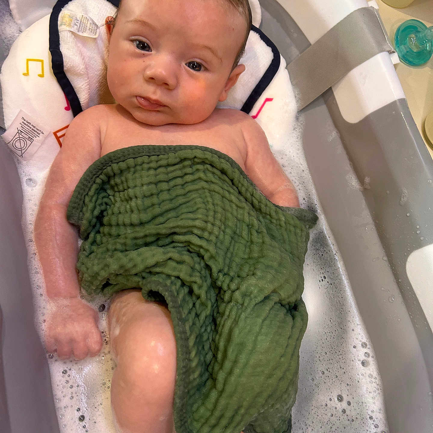 Louis is registered to the contest to win money with this photo: baby, infant, child, bathtub, bath, bubbles, towel, green_towel, wet_hair, face, eyes, portrait, indoors, bathroom, soap, pacifier, water, relaxed, sitting, photography