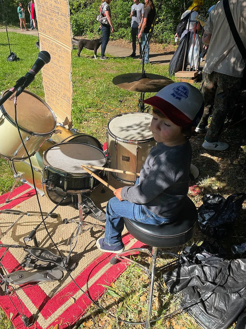 Silas is registered to the contest to win money with this photo: chair, drum, drumhead, drummer, event, grass, hat, headwear, idiophone, leisure, mammal, membranophone, musical_instrument, musician, percussion, percussionist, person, plant, recreation, toddler