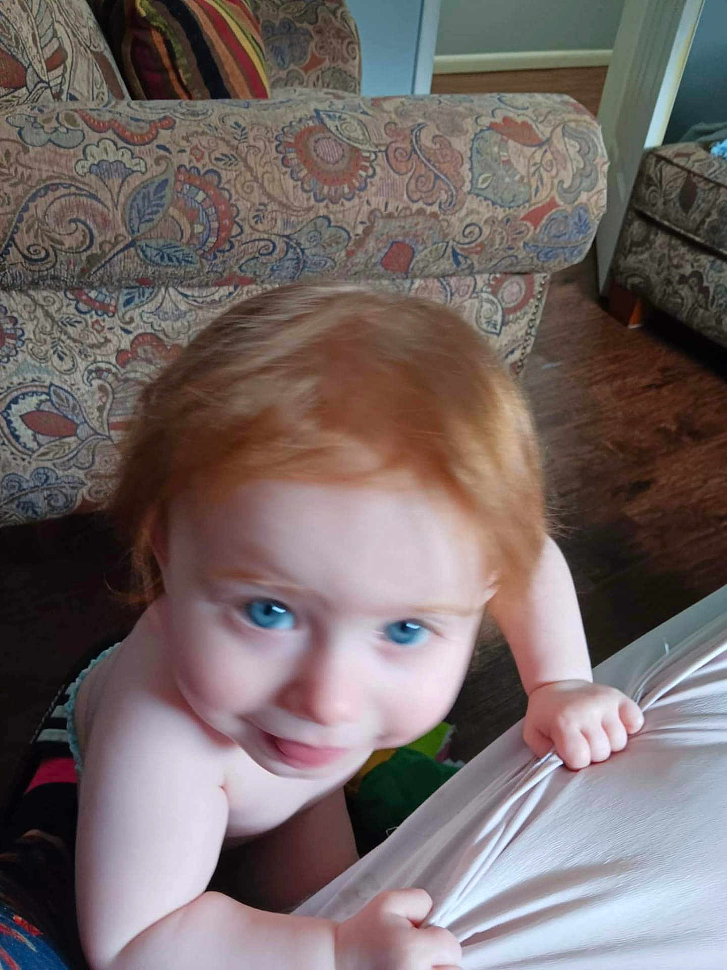 Presley is registered to the contest to win money with this photo: toddler, child, red_hair, blue_eyes, indoor, couch, patterned_fabric, wooden_floor, hand, grip, curious, skin, bare_shoulders, home, carpet, person, furniture, blur, closeup, expression