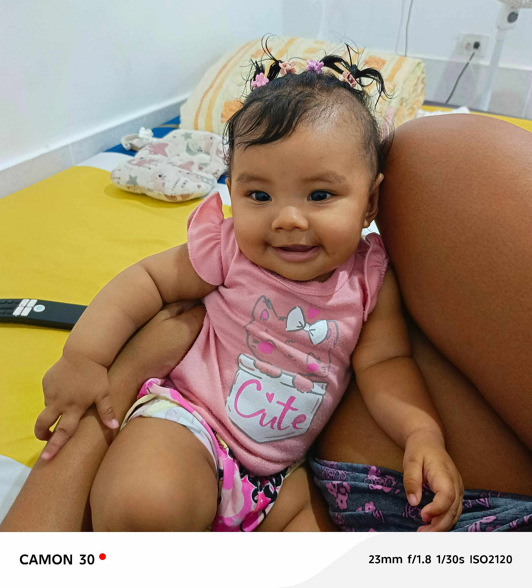 Emma Barreto joined the competition — help win amazing prizes! baby, child, smiling, pink_shirt, cute, hair_clips, indoor, bed, pillow, remote_control, person, skin, legs, hand, happy, infant, casual_clothing, portrait, sitting, closeup