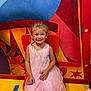 child, girl, pink_dress, smile, orange_chair, colorful_background, playful, happy, indoor, floor, yellow_floor, blue_tape, yellow_tape, sock, small_child, portrait, cute, lacedress, candid, seated