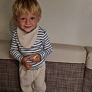 Silas participe au concours pour gagner de l'argent avec cette photo : child, blonde_hair, striped_shirt, pants, bench, cushion, indoor, smile, person, young_child, holding_object, table, casual_clothing, happy, face, head, standing, light, cute, young