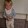 child, blonde_hair, striped_shirt, pants, bench, cushion, indoor, smile, person, young_child, holding_object, table, casual_clothing, happy, face, head, standing, light, cute, young