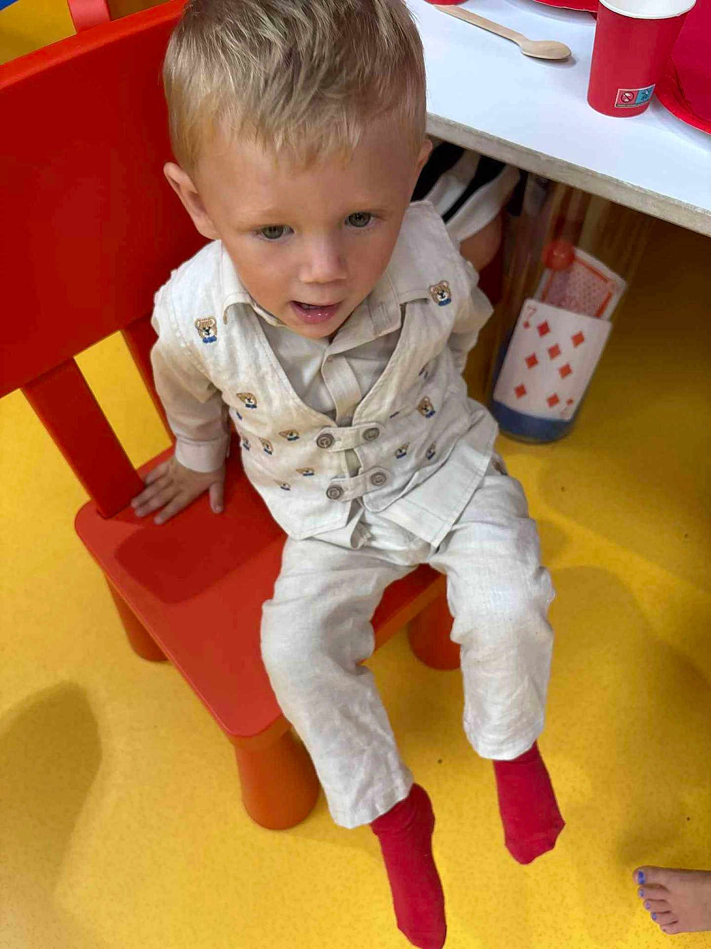 Silas a rejoint le concours — aidez-le/la à gagner de superbes lots ! child, boy, red_chair, yellow_floor, white_outfit, teddy_bear_pattern, red_socks, sitting, indoor, table, playing_cards, red_cup, curious_expression, blond_hair, casual, furniture, floor, person, footwear, clothing