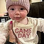 baby, child, face, hat, knit_hat, pom_poms, pink, cream_shirt, game_day, hand, tattoo, nail_polish, smile, indoor, close_up, cute, person, young_child, portrait, apparel