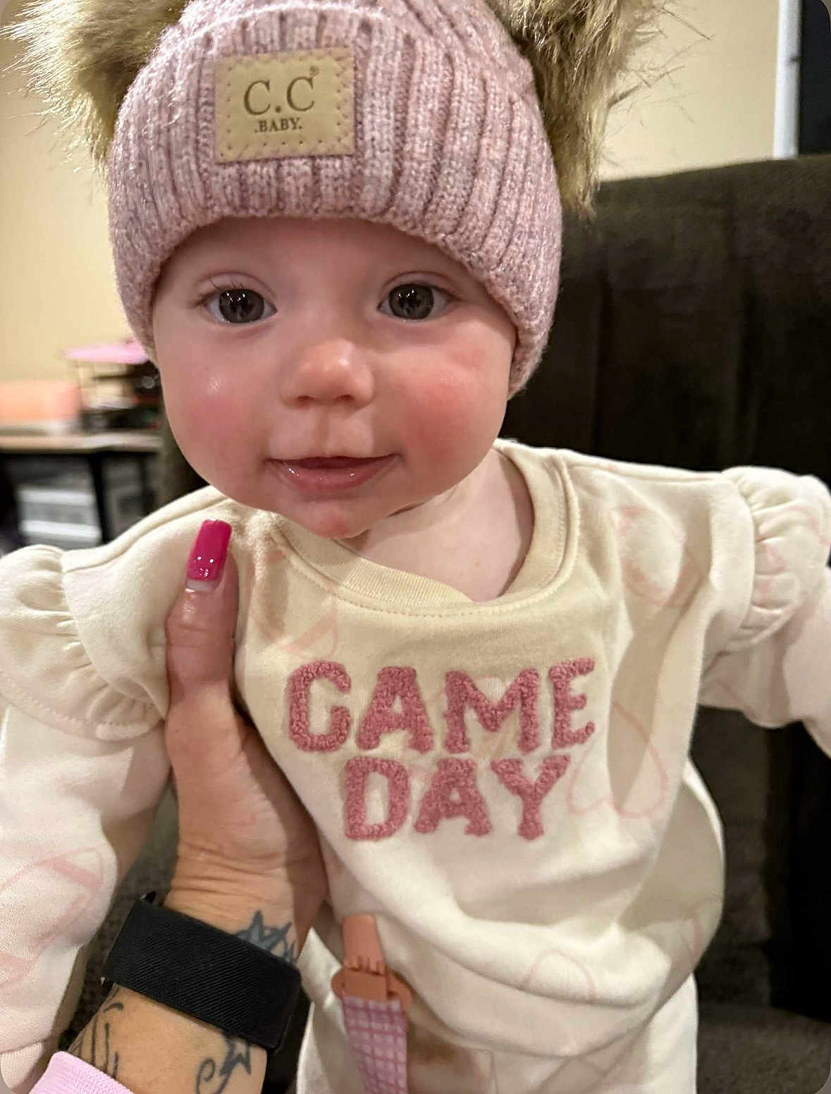 Everly is registered to the contest to win money with this photo: baby, child, face, hat, knit_hat, pom_poms, pink, cream_shirt, game_day, hand, tattoo, nail_polish, smile, indoor, close_up, cute, person, young_child, portrait, apparel