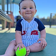Joel is registered to the contest to win money with this photo: baby, child, tennis_ball, outdoor, court, sitting, cute, happy, portrait, sportswear, daylight, playful, person, infant, clothing, green, hand, foot, face, smile