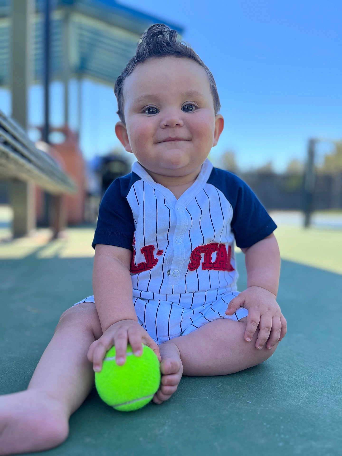 Joel is registered to the contest to win money with this photo: baby, child, tennis_ball, outdoor, court, sitting, cute, happy, portrait, sportswear, daylight, playful, person, infant, clothing, green, hand, foot, face, smile