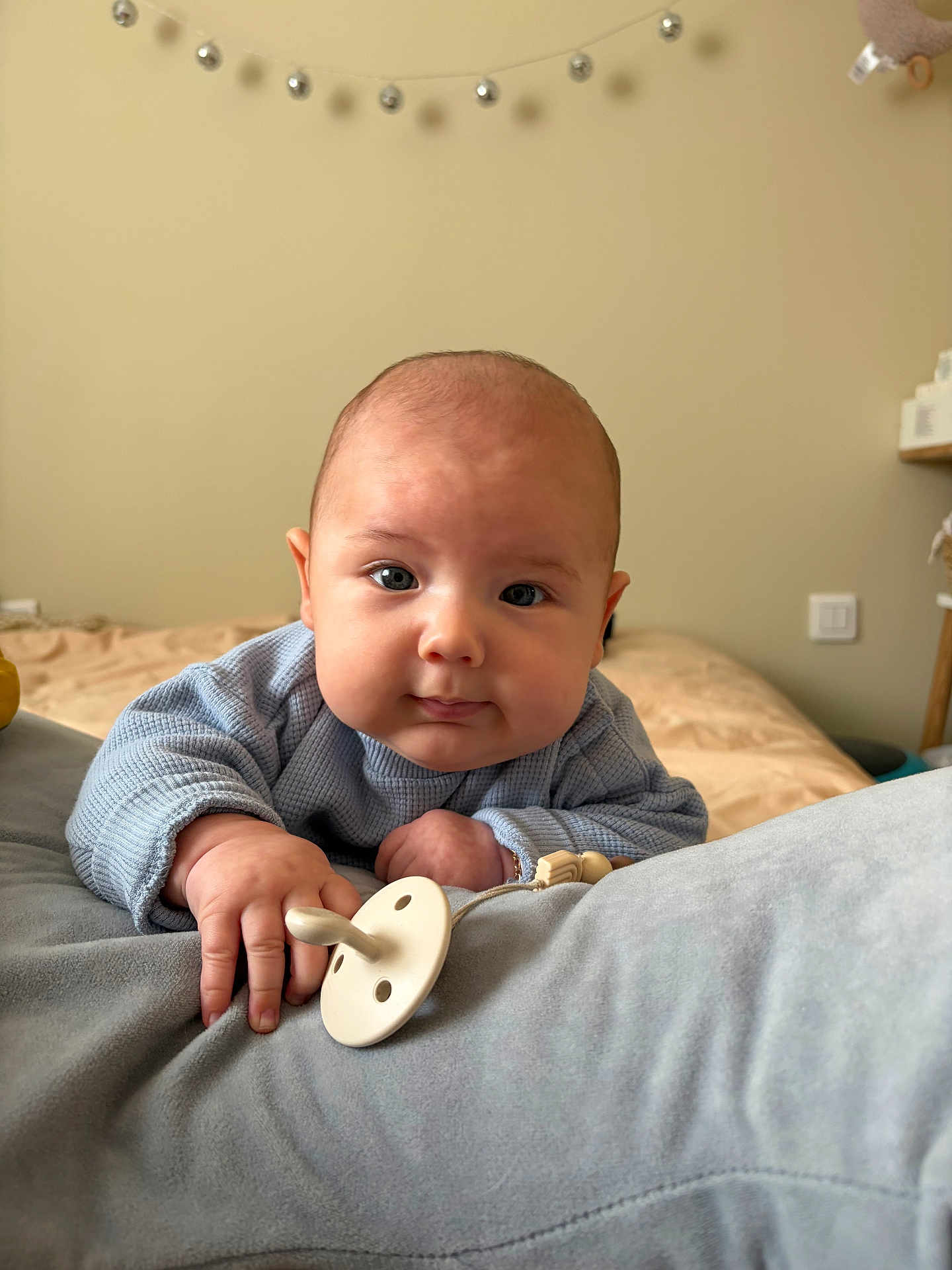 Noa participe au concours pour gagner de l'argent avec cette photo : baby, infant, child, face, eyes, pacifier, hand, sweater, bed, blanket, nursery, wall, string_lights, plush_toy, pillow, chubby_cheeks, close_up, indoor, soft_lighting, looking_at_camera