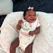 Karielle Eady is registered to the contest to win money with this photo: baby, smiling, infant, pink_bow, bib, onesie, purple_socks, pillow, patterned_pillow, happy, lying_down, indoor, child, cute, fist, face, person, soft_texture, cozy, bed