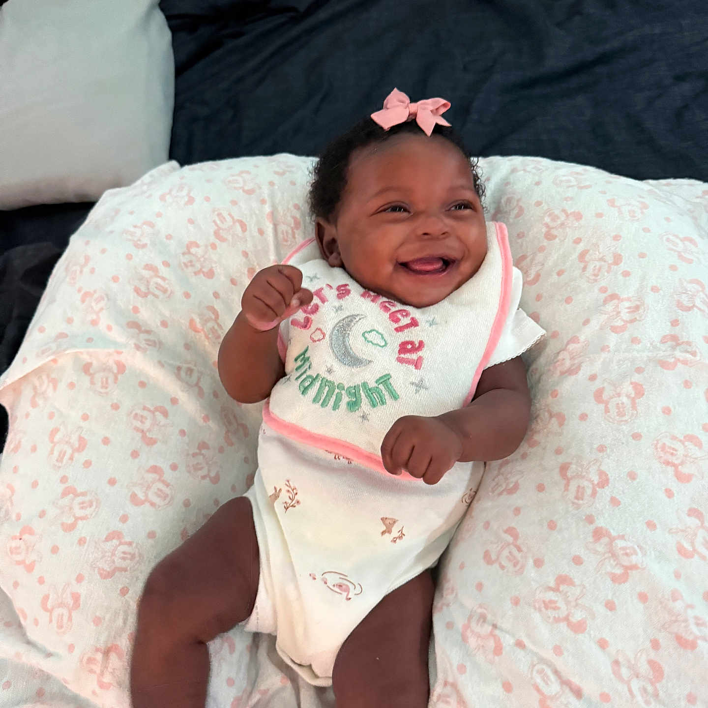 Karielle Eady is registered to the contest to win money with this photo: baby, smiling, infant, pink_bow, bib, onesie, purple_socks, pillow, patterned_pillow, happy, lying_down, indoor, child, cute, fist, face, person, soft_texture, cozy, bed