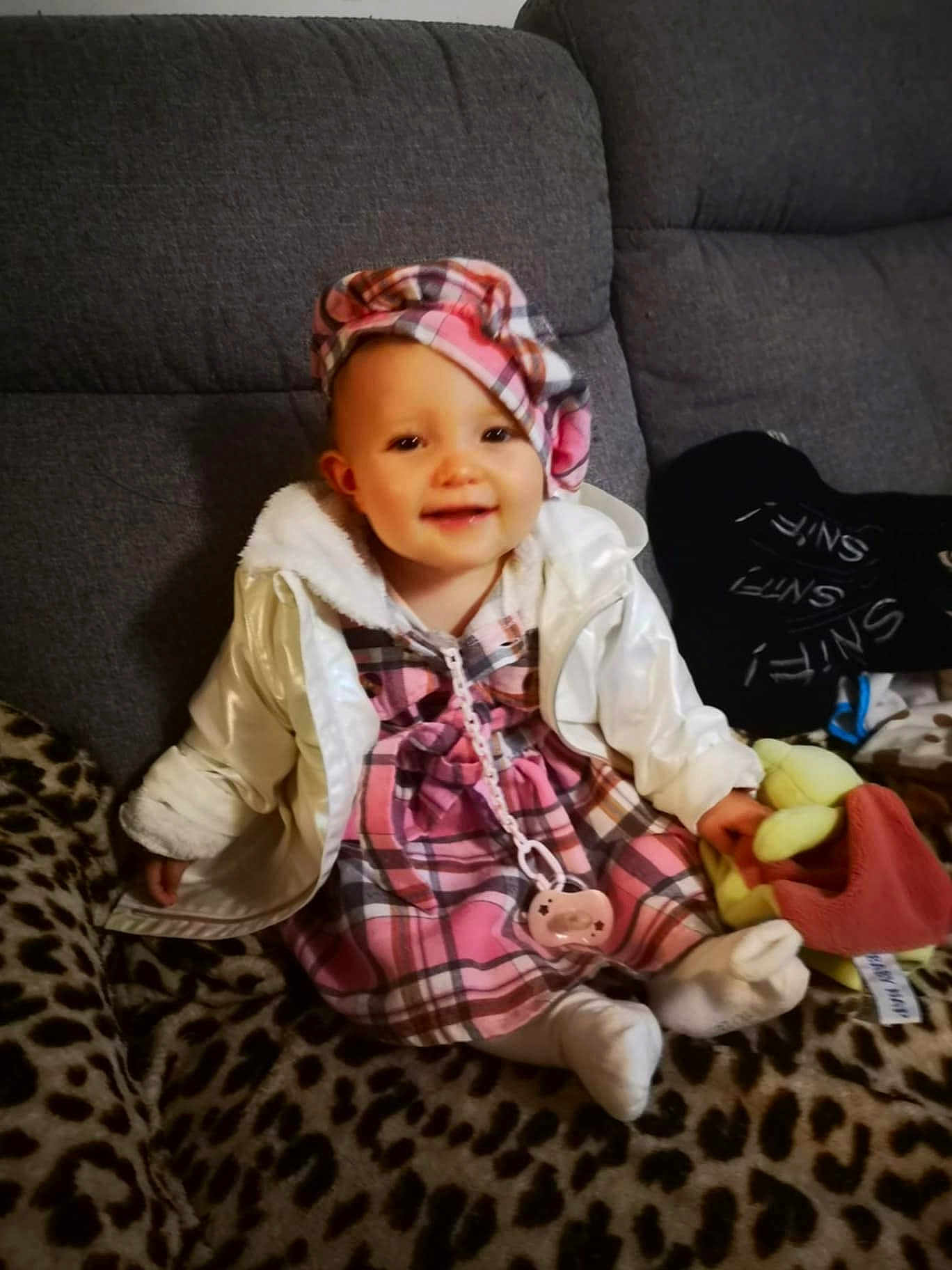 Aëlys participe au concours pour gagner de l'argent avec cette photo : baby, child, smiling, plaid_dress, white_jacket, hat, pacifier, plush_toy, leopard_print, blanket, sitting, indoors, cute, person, face, headwear, cozy, comfort, home, young_child