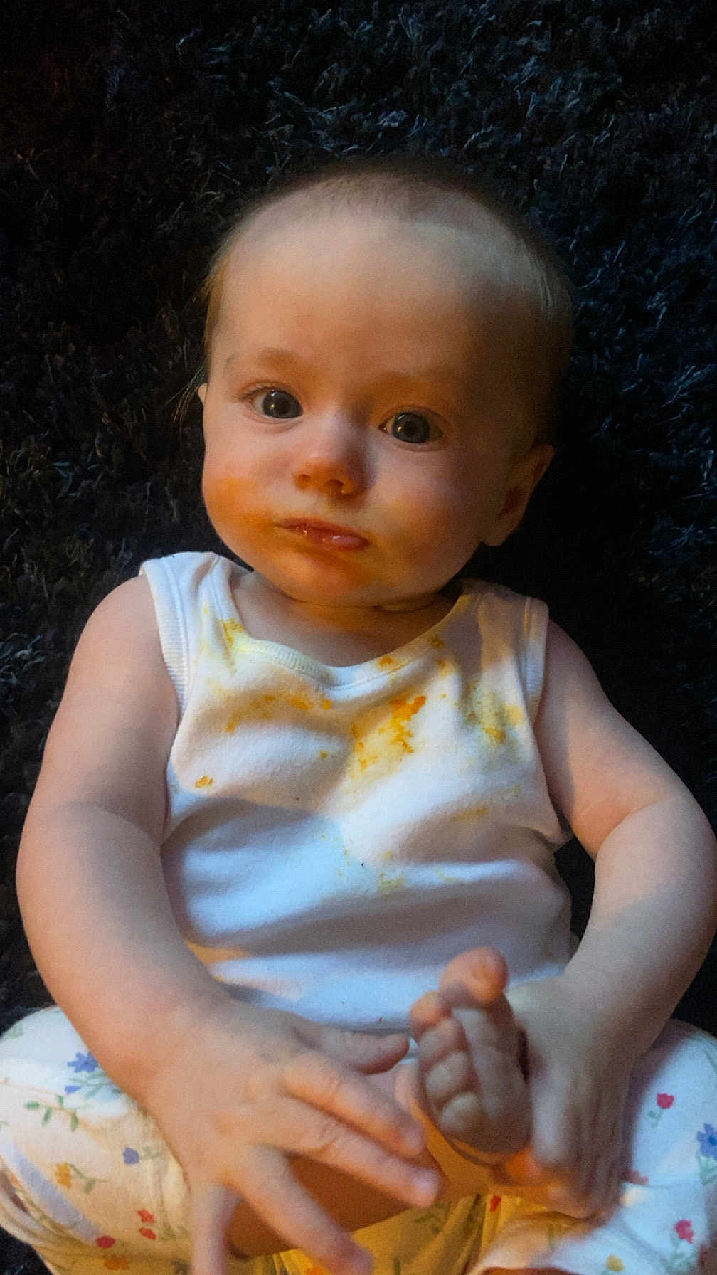 Rosie-May is registered to the contest to win money with this photo: baby, child, infant, face, eyes, white_tank_top, food_stains, hands, feet, floral_pants, sitting, curious, indoors, portrait, cute, expression, skin, person, young, exploration