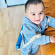 Ares a rejoint le concours — aidez-le/la à gagner de superbes lots ! toddler, child, blue_onesie, backpack, leather_couch, wooden_floor, indoors, smiling, curious, standing, young_child, bright_eyes, cozy_clothing, baby, person, happy, home, casual, cute, portrait