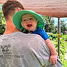 Joey is registered to the contest to win money with this photo: baby, child, adult, green_hat, gray_tshirt, outdoor, shade_cloth, smiling, happy, nature, grass, trees, person, shoulder, sunhat, portrait, daylight, casual_clothing, sign, garden