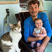 Joey is registered to the contest to win money with this photo: man, baby, cat, chair, wood, window, painting, indoor, smile, happy, person, animal, pet, holding, casual_clothing, green_eyes, baby_bottle, decor, furniture, portrait
